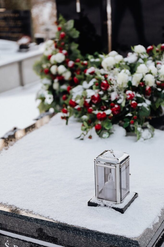 Snow-covered grave with flowers and a candle holder in a winter cemetery setting.