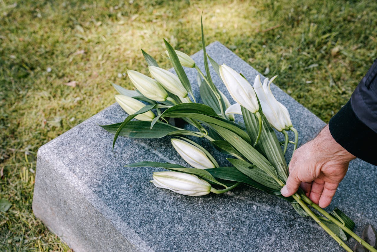 A hand placing white lilies on a granite tombstone in an outdoor cemetery.